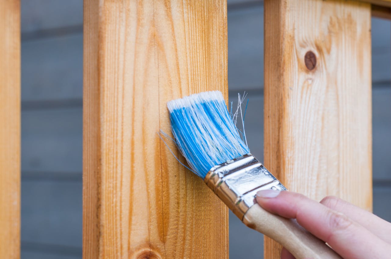 services-03 A hand painting a wooden fence plank with a blue paintbrush outdoors. Perfect for DIY and renovation themes.