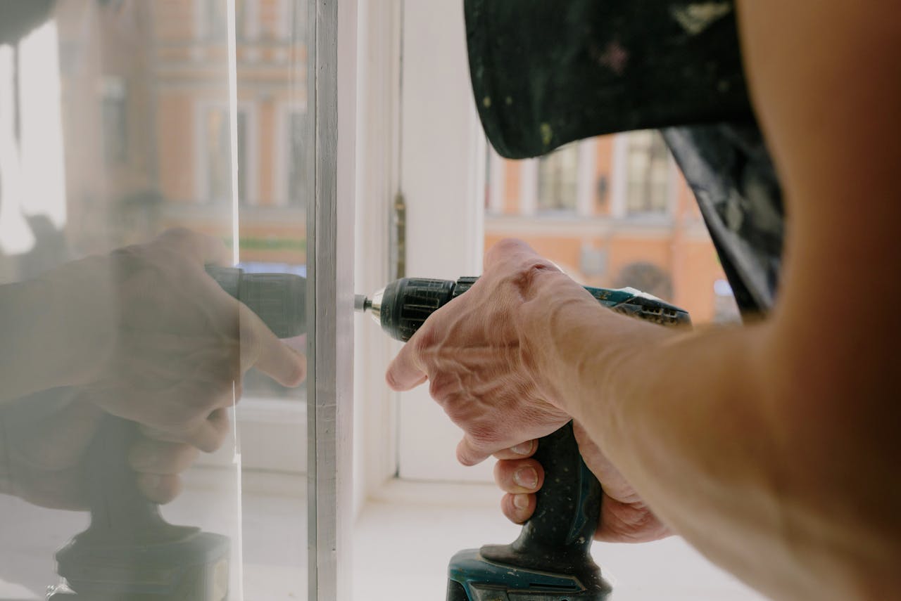 services-04 Close-up of a handyman's hands using a drill to fix a window inside an apartment.